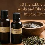 A bottle of natural Amla and Bhringraj hair oil sitting next to fresh Indian Gooseberries and green herbs on a wooden table.