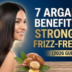 A bottle of golden argan hair oil sitting next to raw argan kernels on a smooth wooden surface.