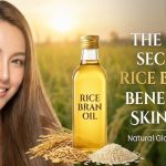 A wooden spoon filled with golden rice bran oil sitting next to a bowl of rice grains and a bottle of hair oil.